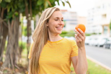 Açık havada elinde portakal tutan genç sarışın kadın. Mutlu bir ifadeyle.