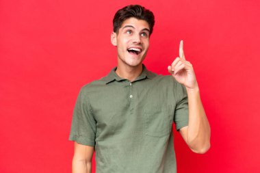 Genç, beyaz, yakışıklı, kırmızı arka planda izole edilmiş bir adam. Çözümü bulmak için parmağını kaldırıyor.