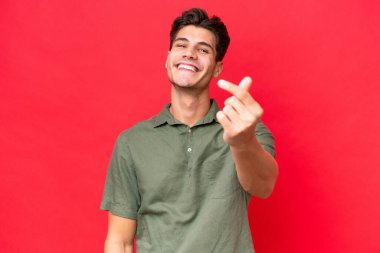 Kafkasyalı yakışıklı genç adam kırmızı arka planda izole edilmiş, para hareketi yapıyor.