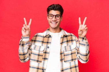 Kırmızı arka planda izole edilmiş beyaz, yakışıklı genç bir adam zafer işaretini iki elle gösteriyor.