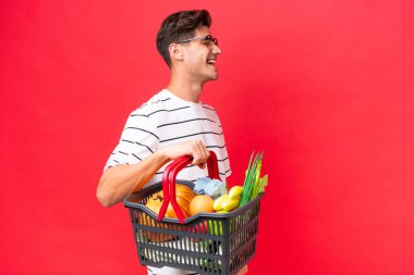 Kafkasyalı genç adam elinde yiyecek dolu bir sepet tutuyor. Kırmızı arka planda izole edilmiş. Yan yana gülüyor.