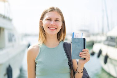 Sarışın genç bir kadın elinde pasaport, dışarıda gülümsüyor.
