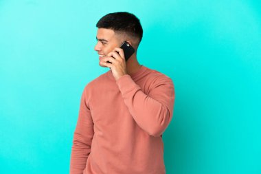Soyutlanmış mavi arka planda genç yakışıklı bir adam cep telefonuyla sohbet ediyor.