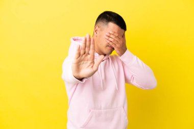 Soyutlanmış sarı arka planda genç yakışıklı adam jesti yapıyor ve yüzünü kapatıyor.