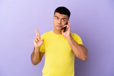 İzole edilmiş mor arka planda cep telefonu kullanan genç ve yakışıklı bir adam parmaklarını çapraz yapıp en iyisini diliyor.