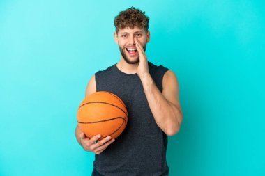 Basketbol oynayan yakışıklı genç adam mavi arka planda izole edilmiş ve ağzı açık bağırıyor.