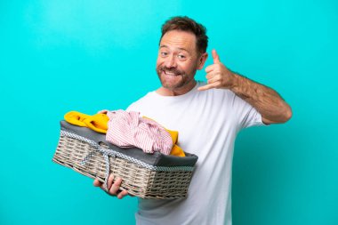 Mavi arka planda izole edilmiş çamaşır sepetini tutan orta yaşlı bir kadın telefon jesti yapıyor. Bana geri dönüş işareti ver.