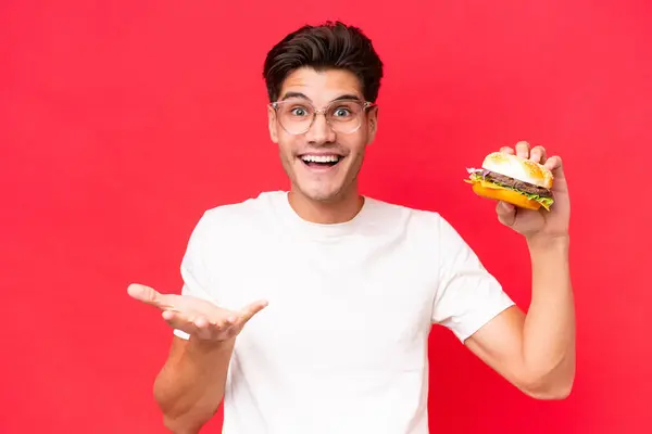 Młody Biały Mężczyzna Trzymając Hamburgera Odizolowany Czerwonym Tle Wstrząśniętym Wyrazem — Zdjęcie stockowe
