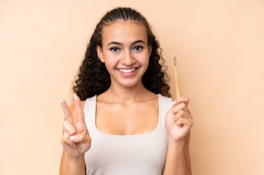 Bej arka planda dişlerini fırçalayan genç bir kadın gülümsüyor ve zafer işareti gösteriyor.