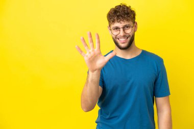 Sarı arka planda izole edilmiş yakışıklı beyaz adam parmaklarıyla beşi sayıyor.