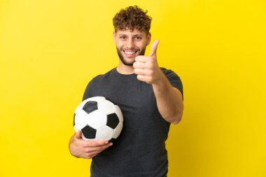 Yakışıklı genç futbolcu sarı arka planda izole edildi. Başparmakları havada çünkü iyi bir şey oldu.