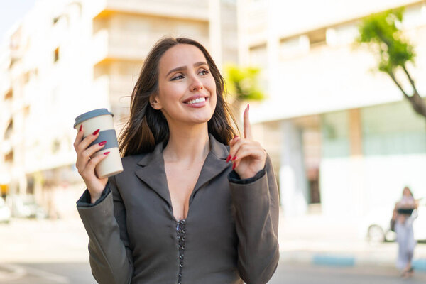Young woman holding a take away coffee at outdoors intending to realizes the solution while lifting a finger up