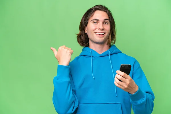 年轻英俊男子用手机用绿色背景隔离 并指向侧边 — 图库照片