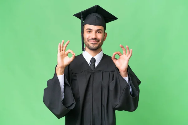 Молодий Випускник Університету Над Ізольованим Тлом Показує Знак Пальцями — стокове фото