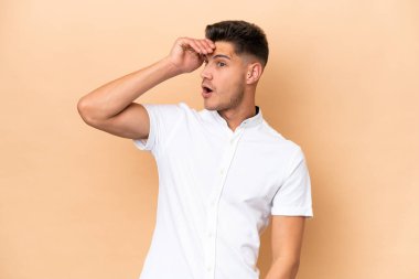 Beyazlı genç adam bej arka planda izole edilmiş. Yan tarafa bakarken sürpriz jest yapıyor.
