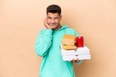 Elinde fast food tutan beyaz bir adam bej arka planda izole edilmiş hayal kırıklığına uğramış ve kulaklarını kapatmış.