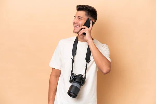 年轻的摄影师高加索人男子孤立在米黄色背景下与手机保持对话 — 图库照片