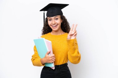 Arap üniversitesinden mezun bir kadın elinde beyaz arka planda kitap tutuyor gülümsüyor ve zafer işareti gösteriyor.