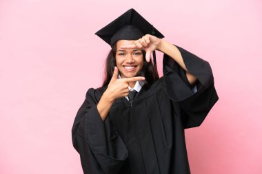 Üniversiteden mezun genç bir Kolombiyalı kadın pembe arka planda izole edilmiş, odaklanmış bir yüzle. Çerçeve sembolü