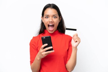 Genç Kolombiyalı bir kadın beyaz arka planda izole edilmiş cep telefonuyla alışveriş yapıyor ve elinde şaşırmış bir ifadeyle bir kredi kartı tutuyor.