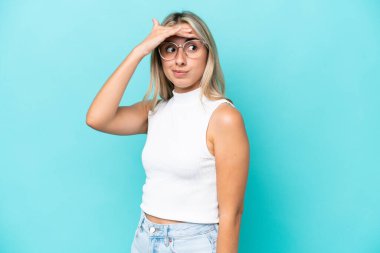 Mavi arka planda izole edilmiş genç beyaz kadın yan tarafa bakarken sürpriz jest yapıyor.