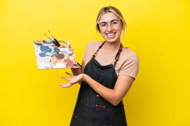 Sarı arka planda yalıtılmış bir palet tutan beyaz bir kadın davet etmek için ellerini yana uzatıyor.