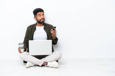 Yerde dizüstü bilgisayarı olan genç Brezilyalı bir adam beyaz üzerine izole bir şekilde oturmuş elinde kahve ve bir yandan da bir şeyler düşünürken bir yandan da cep telefonu tutuyordu.