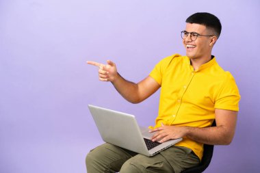 Genç bir adam dizüstü bilgisayarla bir koltukta oturuyor ve bir ürünü gösteriyor.