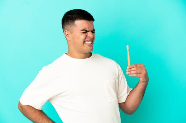 Genç ve yakışıklı bir adam dişlerini fırçalıyor. Mavi arka plan üzerinde. Çabaladığı için sırt ağrısı çekiyor.