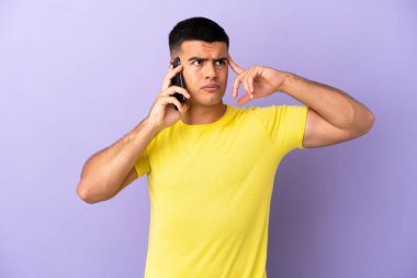 İzole edilmiş mor arka planda cep telefonu kullanan genç yakışıklı bir adam. Şüpheleri ve düşünceleri var.