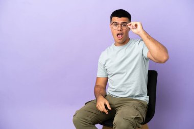 Sandalyede oturan genç bir adam izole edilmiş arka planda gözlüklü ve şaşırmış.