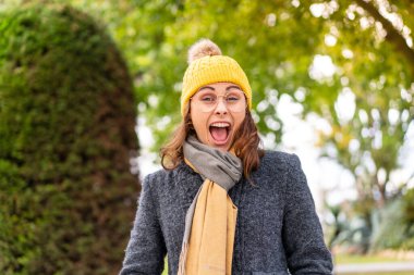 Açık havada esmer bir kadın kışlık ceket giyiyor ve yüzünde sürpriz bir ifade var.