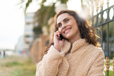 Açık havada genç beyaz bir kadın cep telefonuyla sohbet ediyor.