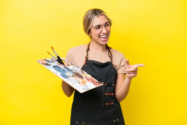黄色の背景に指を向け 製品を提示するパレットを保持若い芸術家の白人女性 — ストック写真