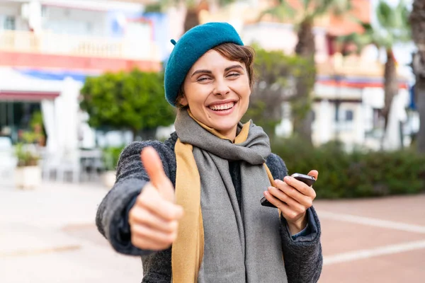 親指を立てながら屋外で携帯電話を使用しているブルネットの女性 — ストック写真