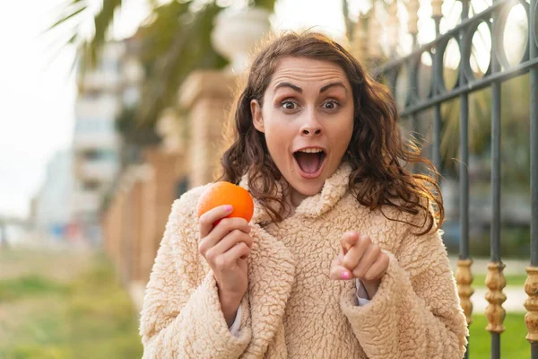 Mladá Běloška Drží Pomeranč Venku Překvapen Ukazuje Dopředu — Stock fotografie