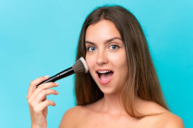 Young caucasian woman isolated on blue background holding makeup brush and surprised