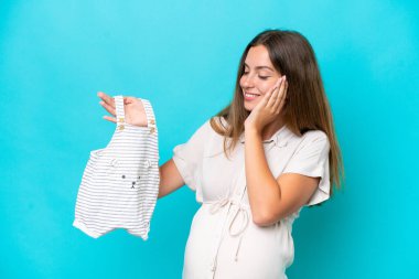 Young caucasian woman isolated on blue background pregnant and holding baby clothes