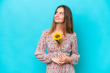 Kafkasyalı genç bir kadın elinde ayçiçeği tutarken mavi arka planda izole bir şekilde yukarı bakarken bir fikir düşünüyor.