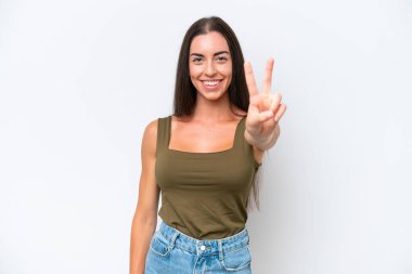 Beyaz arka planda izole edilmiş genç beyaz kadın gülümsüyor ve zafer işareti gösteriyor.