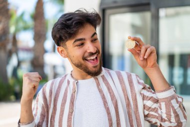 Yakışıklı bir Arap elinde Bitcoin ile dışarıda zaferi kutluyor.