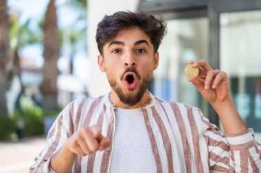 Açık havada Bitcoin tutan yakışıklı bir Arap şaşırmış ve ön tarafı işaret ediyor.