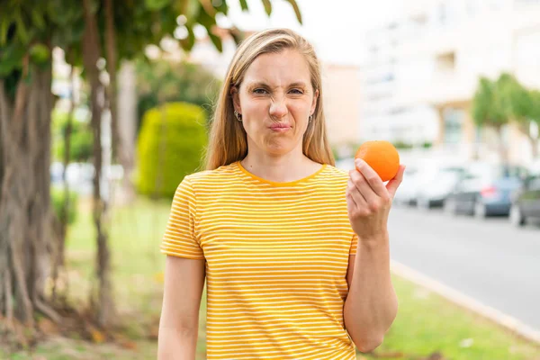 Junge Blonde Frau Hält Draußen Eine Orange Mit Traurigem Gesichtsausdruck — Stockfoto