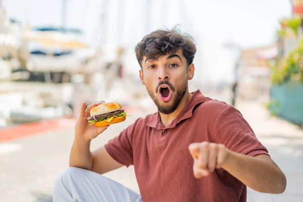 Jonge Arabische Knappe Man Houdt Een Hamburger Buiten Verrast Wijst — Stockfoto