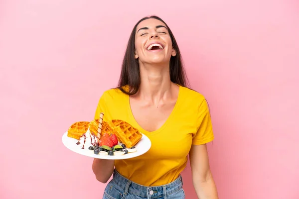 年轻的高加索女人拿着用粉色背景笑隔离的华夫饼 — 图库照片