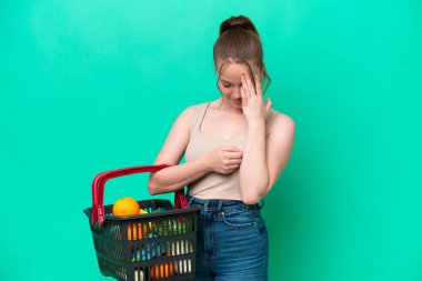 Elinde yiyecek dolu bir sepet taşıyan genç bir kadın. Yeşil arka planda, yorgun ve hasta bir ifadeyle izole edilmiş.