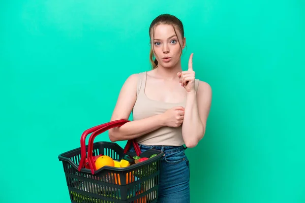 指を持ち上げながら解決策を実現しようと緑の背景に隔離された食べ物でいっぱいの買い物かごを持っている若い女性 — ストック写真