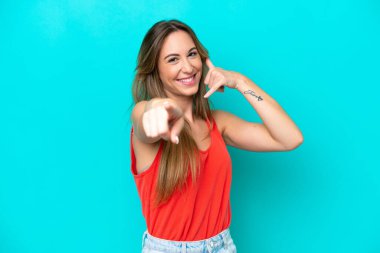 Mavi arka planda izole edilmiş genç beyaz kadın telefon hareketlerinde bulunuyor ve ön tarafı işaret ediyor.