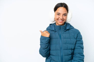 Young hispanic woman wearing a winter earmuffs isolated on white background pointing to the side to present a product