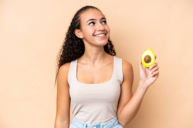 Elinde bej arka planda izole edilmiş avokado tutan genç bir kadın gülümserken yukarı bakıyor.